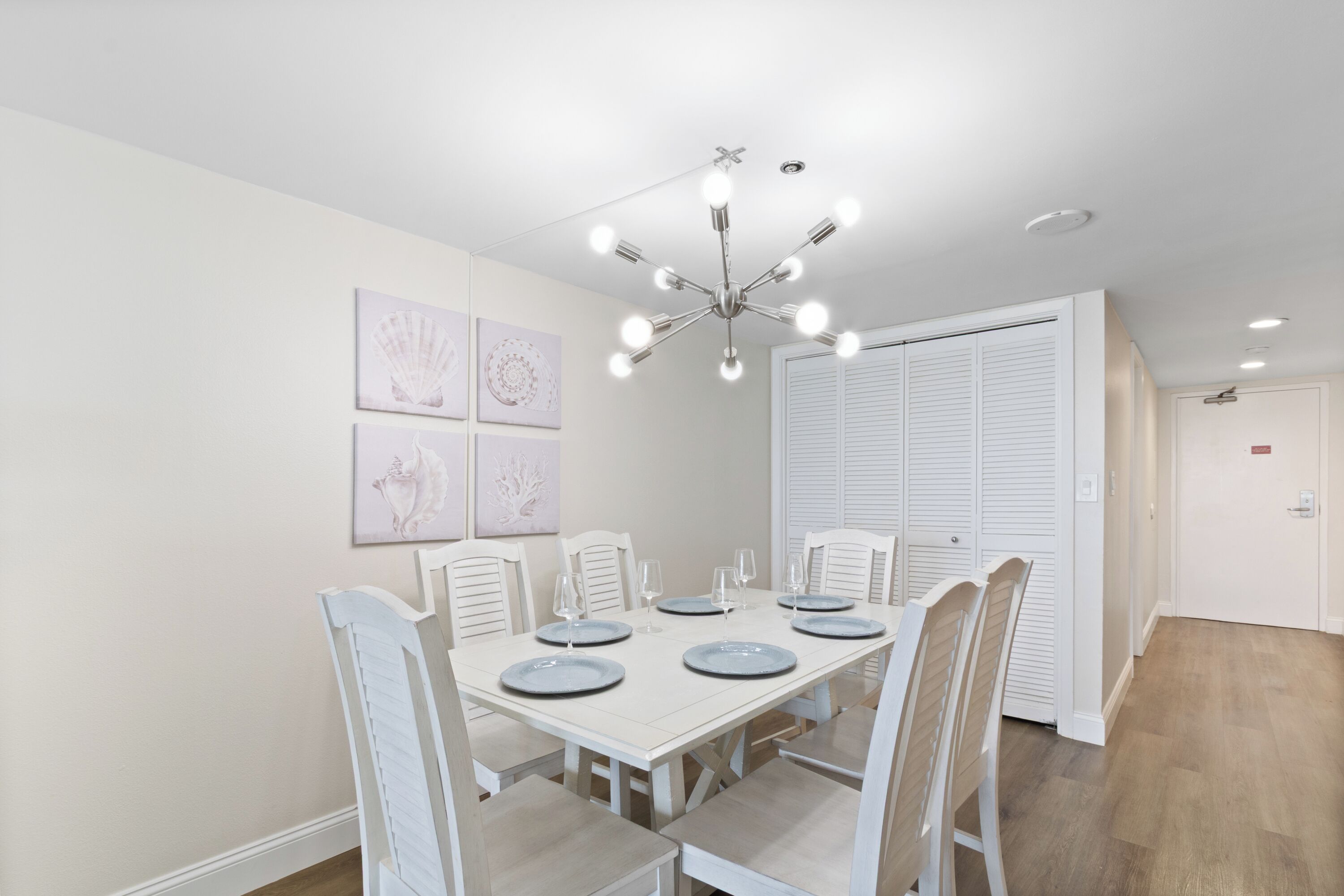 A dining room with a white table set for six, white chairs, a modern chandelier, and a white door in the background.