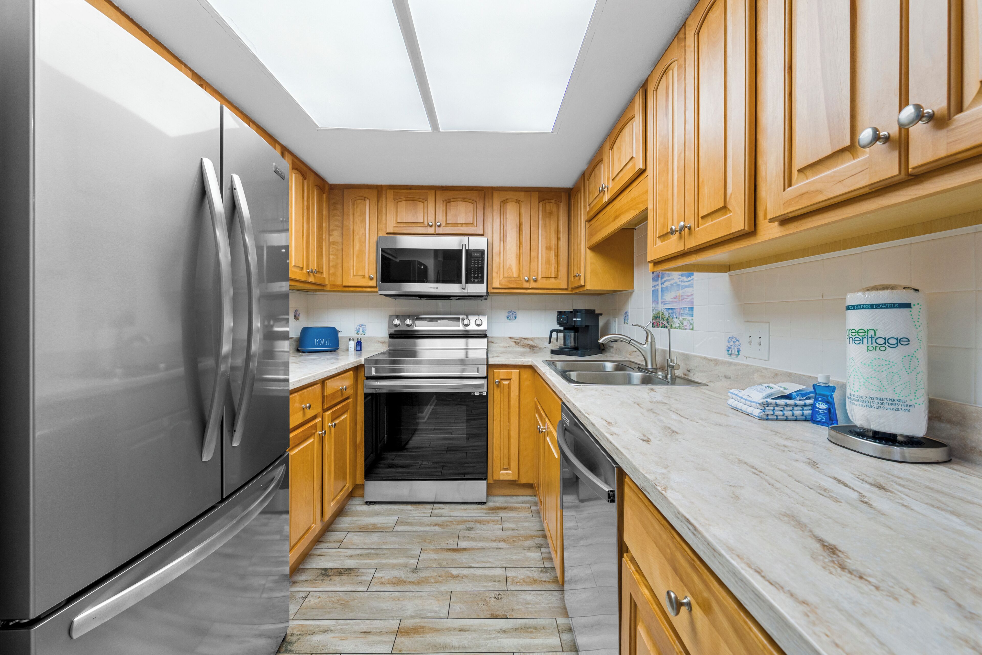 A modern kitchen with stainless steel appliances, wooden cabinets, a skylight, and a marble countertop.