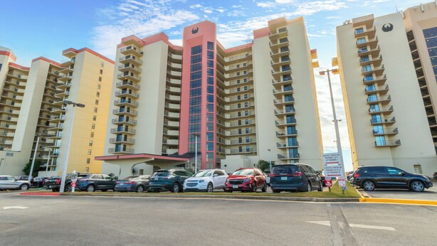 A multi-story residential building with balconies, a parking lot with several cars, and a clear sky.