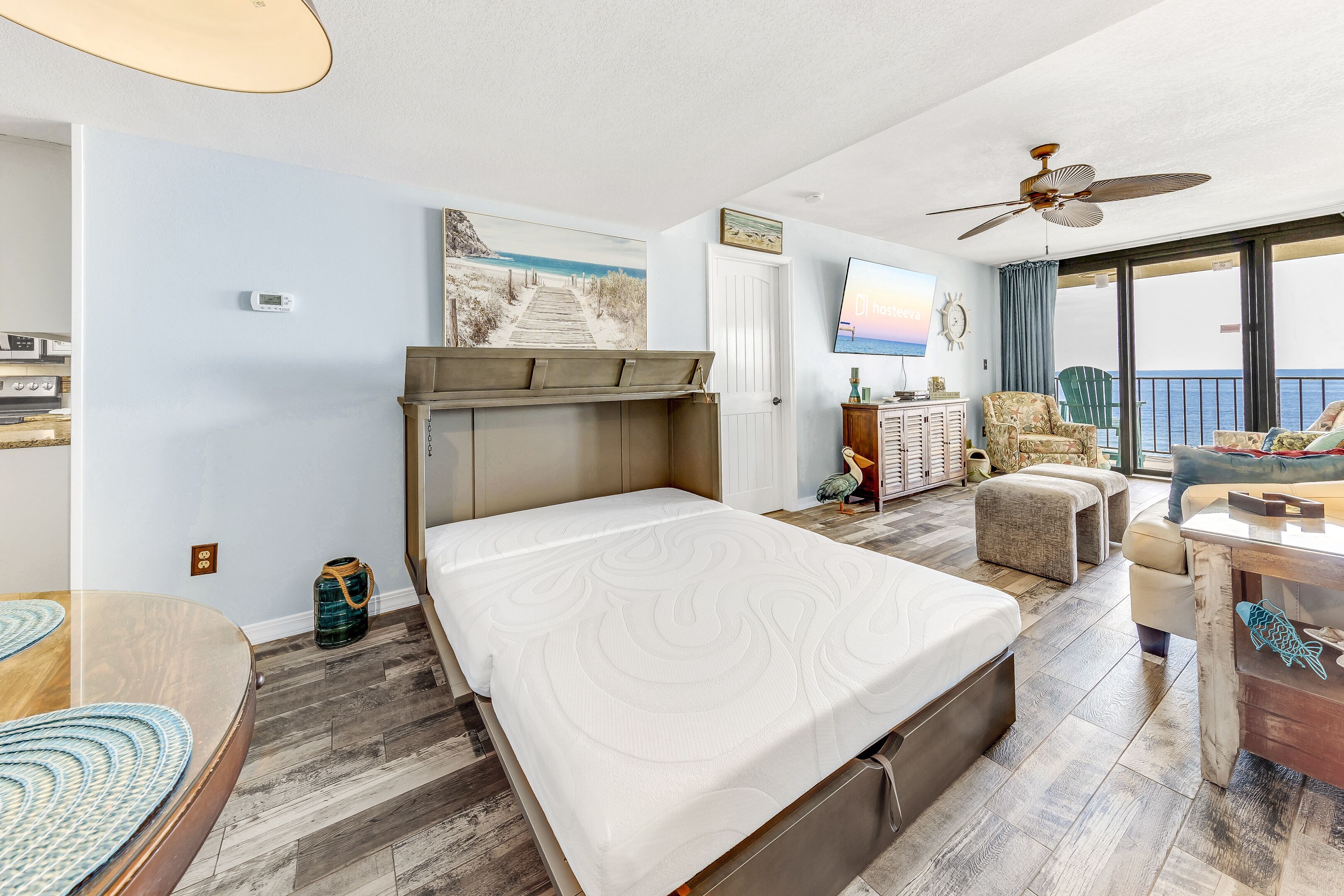 A hotel room with a bed, a desk, a chair, a balcony with a view, and a ceiling fan.