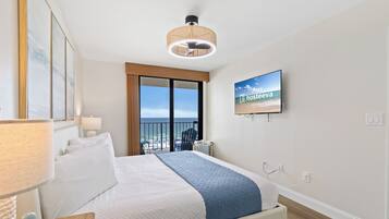 A hotel room with a bed, a television, a view of the beach, and a balcony.