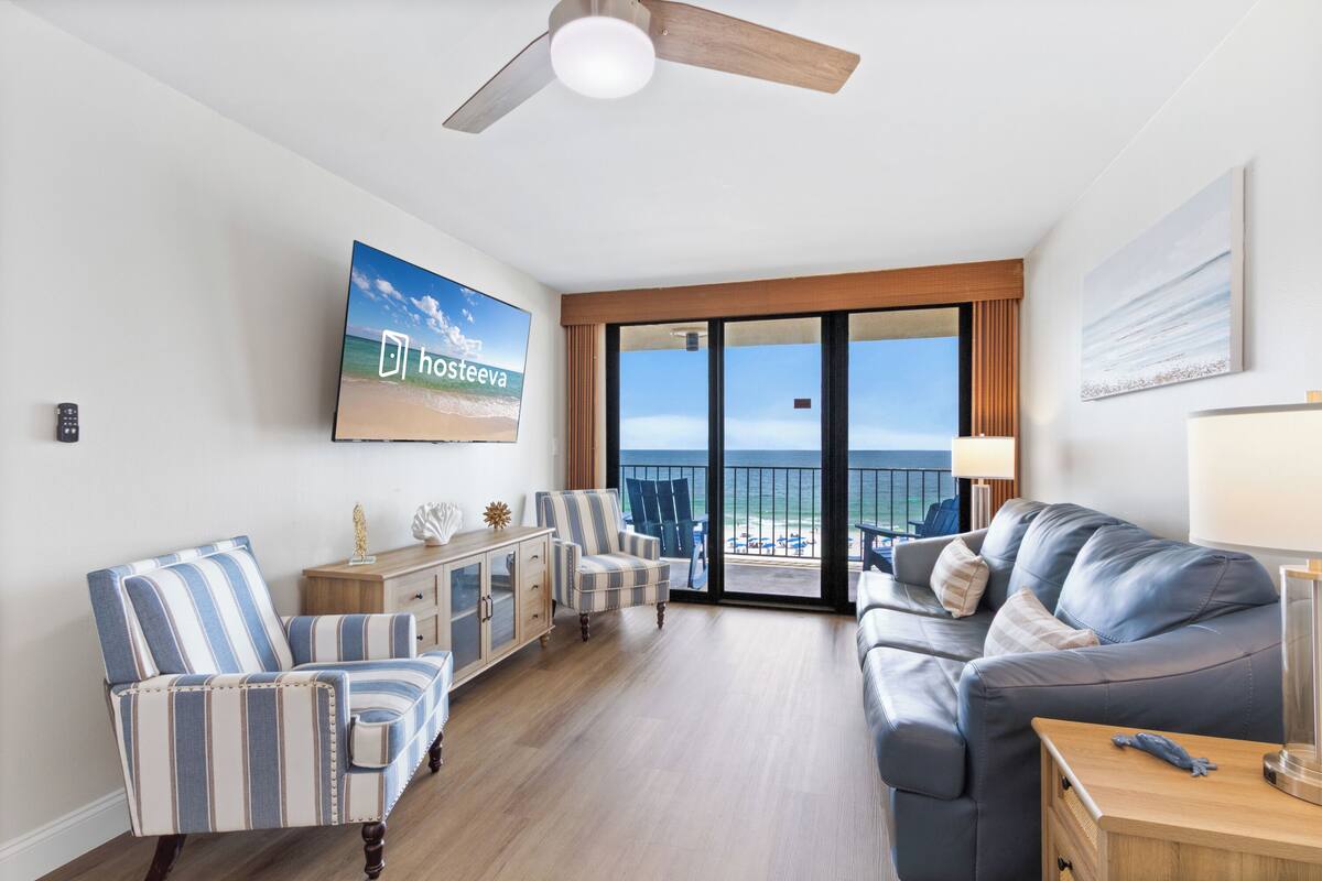 A living room with a sofa, striped armchairs, a cabinet, a television, and a balcony with a view of the ocean.