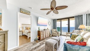 A spacious living room with a large window, a ceiling fan, a television, and a balcony with a view of the ocean.