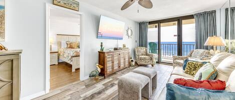 Un salón amplio con un ventanal, ventilador de techo, televisor y balcón con vista al océano.