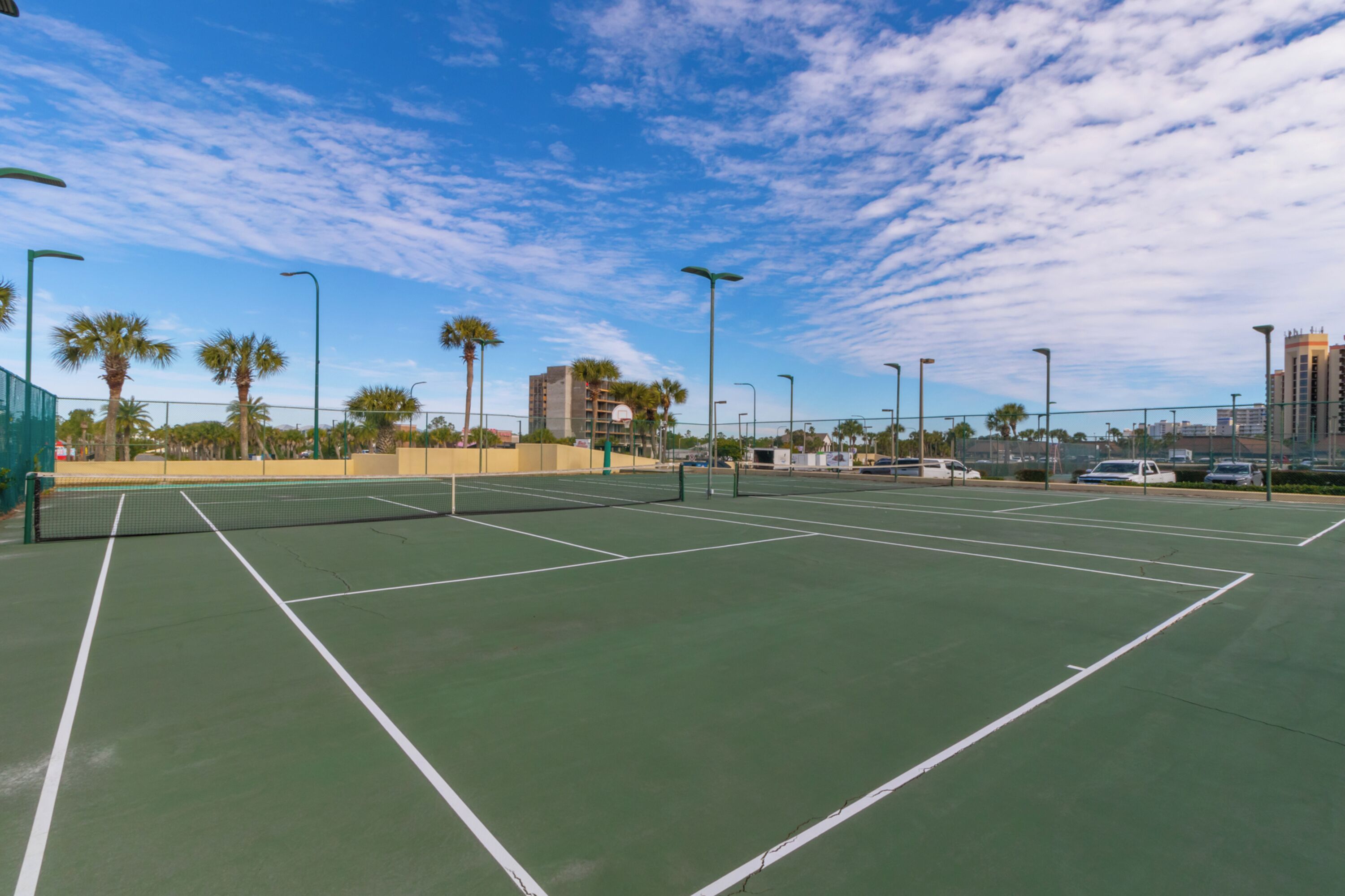 An outdoor tennis court with multiple courts, surrounded by palm trees and street lamps, with buildings in the background.