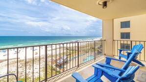 A balcony with a view of the beach and ocean, blue chairs, and a pool area.