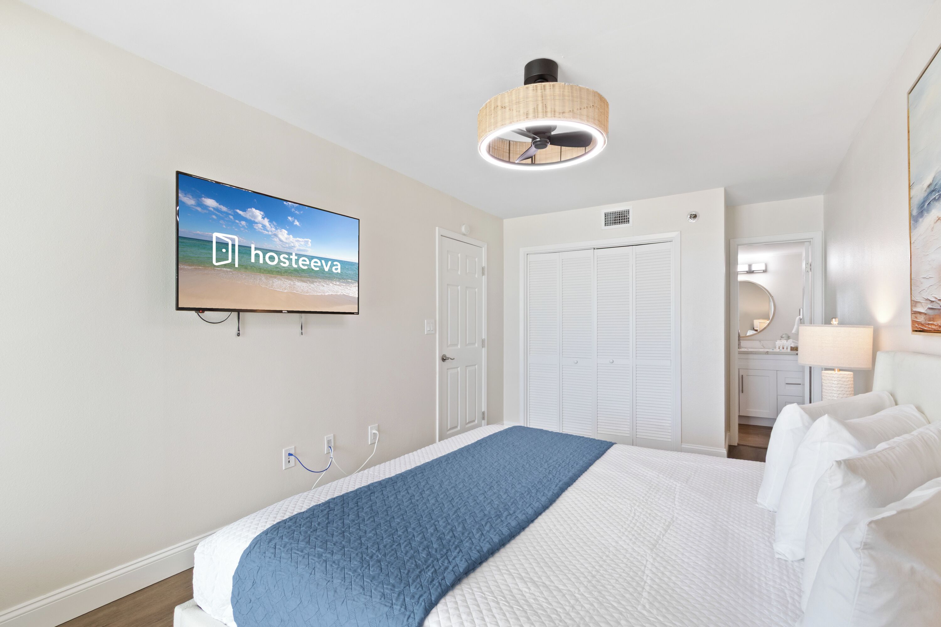 Ein Hotelzimmer mit einem Bett, einem Fernseher, der den Namen des Hotels zeigt, einem Deckenventilator und einer Tür zu einem Badezimmer.