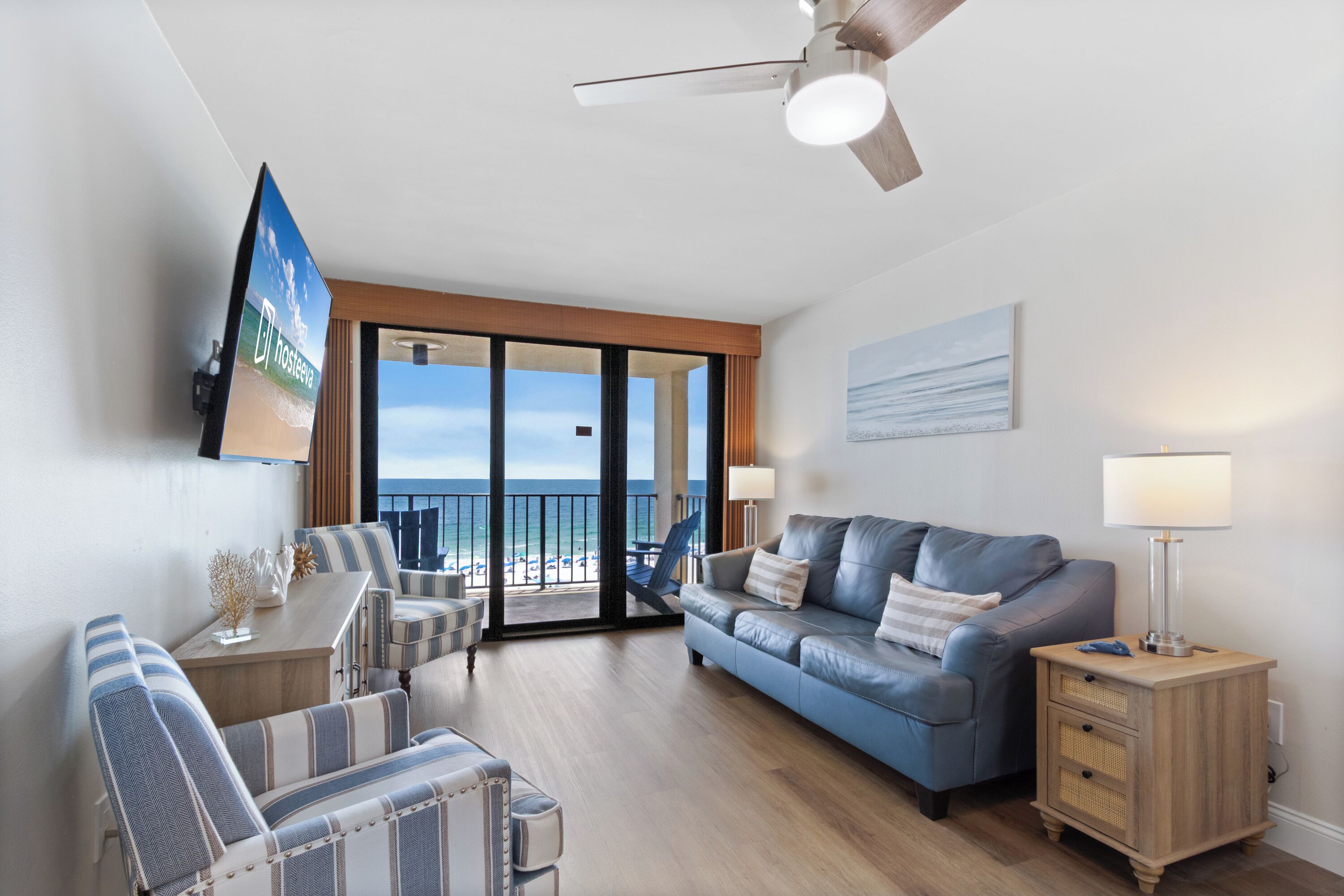 A modern living room with a sofa, armchairs, a coffee table, a TV, a ceiling fan, and a view of the ocean through large glass doors.