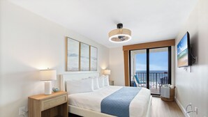 A hotel room with a bed, a nightstand, a lamp, a television, and a view of the beach.