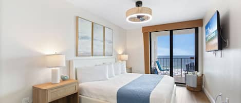 A hotel room with a bed, a nightstand, a lamp, a television, and a view of the beach.