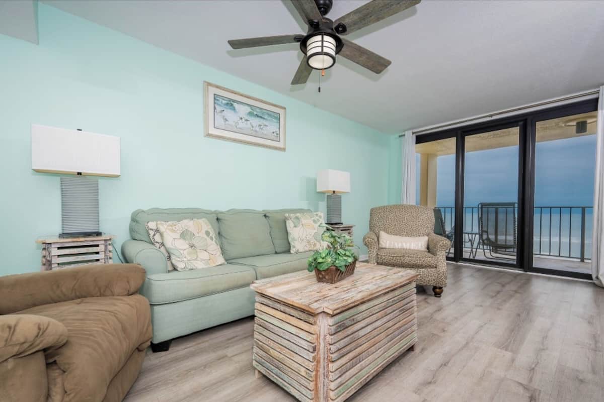 A living room with a sofa, a chair, a coffee table, a ceiling fan, a lamp, a framed picture, and a view of the ocean through large glass doors.