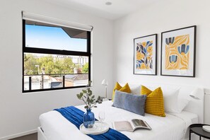 A hotel room with a large window, a bed with white bedding, a blue vase with flowers, a book, and two framed artworks on the wall. - Caulfield Elegance: Chic Abode (Caulfield North)