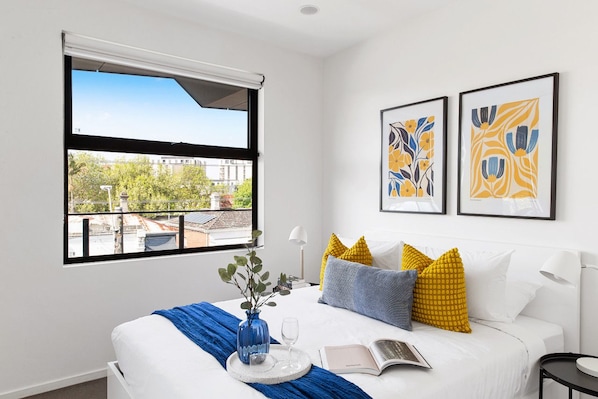A hotel room with a large window, a bed with white bedding, a blue vase with flowers, a book, and two framed artworks on the wall.