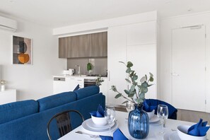 A modern dining area with a blue sofa, a white table set for two, and a kitchen in the background. - Caulfield Elegance: Chic Abode (Caulfield North)