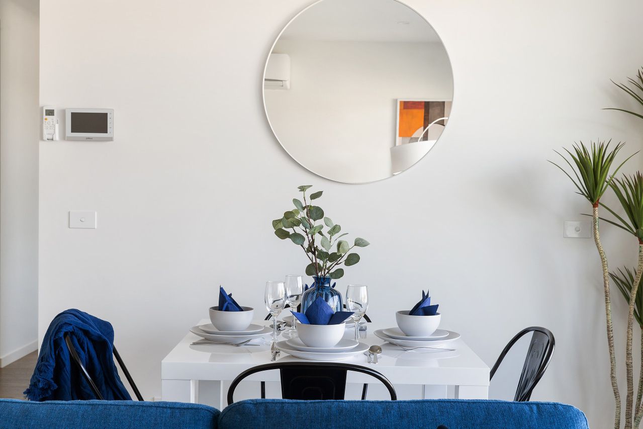 A dining area with a white table set for four, a round mirror above, and a blue vase with green foliage.