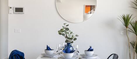Zona pranzo con tavolo bianco apparecchiato per quattro persone, uno specchio rotondo sopra e un vaso blu con verde decorazione.