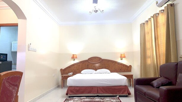 A hotel room with a wooden headboard, two bedside lamps, a bed with white linens, a brown leather armchair, and a patterned rug. - Super OYO 151 Manam 2 Hotel Apartment (Muscat)
