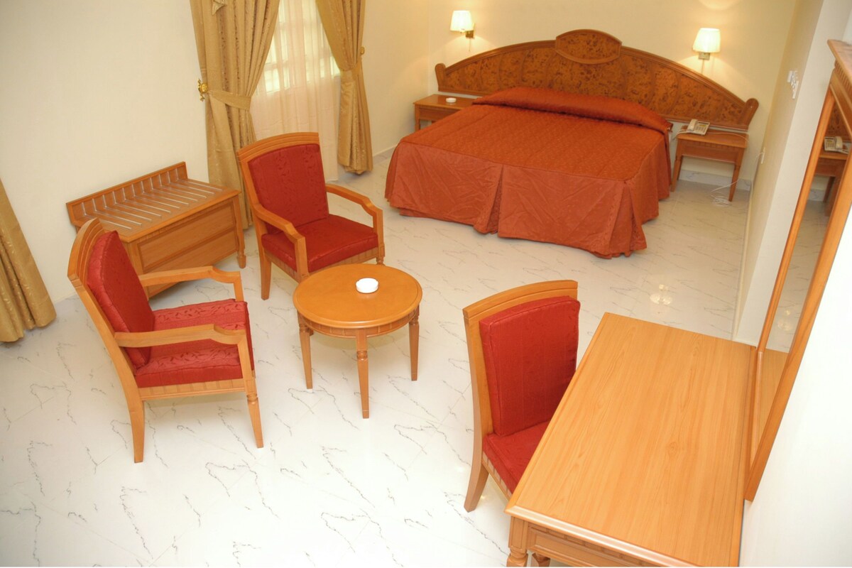A hotel room with a bed, two armchairs, a small table, and a wooden bench.