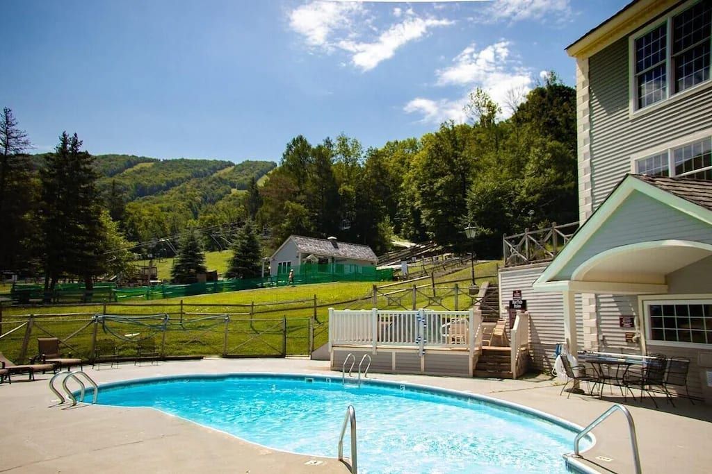 A swimming pool with a wooden deck, a fenced area, and a house in the background.