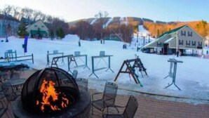 Área externa coberta de neve com lareira, mesas e cadeiras, sugerindo um ambiente de resort de inverno.