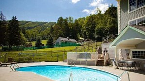 Piscina com deck de madeira, área cercada e uma casa ao fundo.