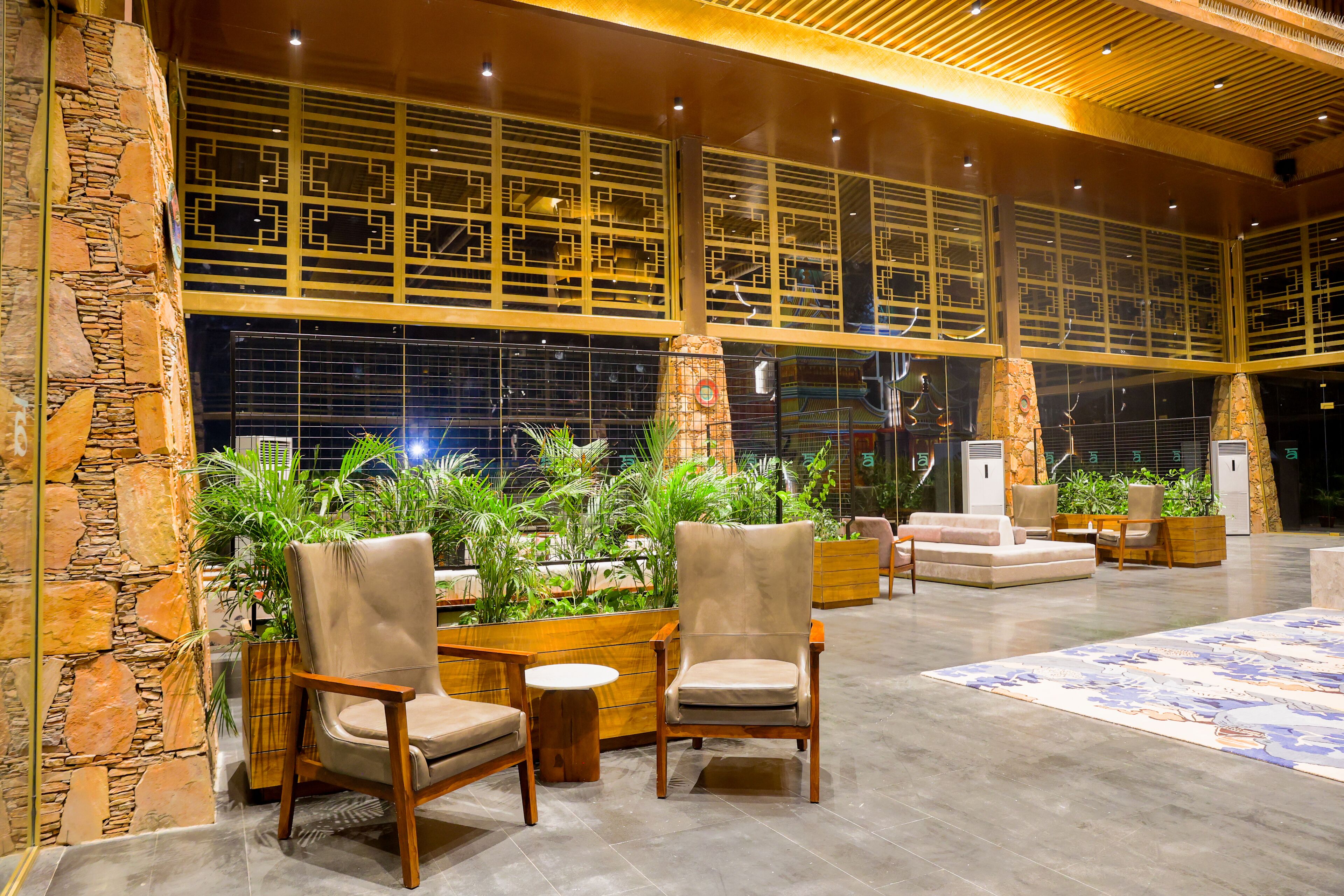 A spacious lobby with a high ceiling, stone walls, and large windows with decorative grills. There are several seating areas with chairs and tables, and a variety of plants are visible.