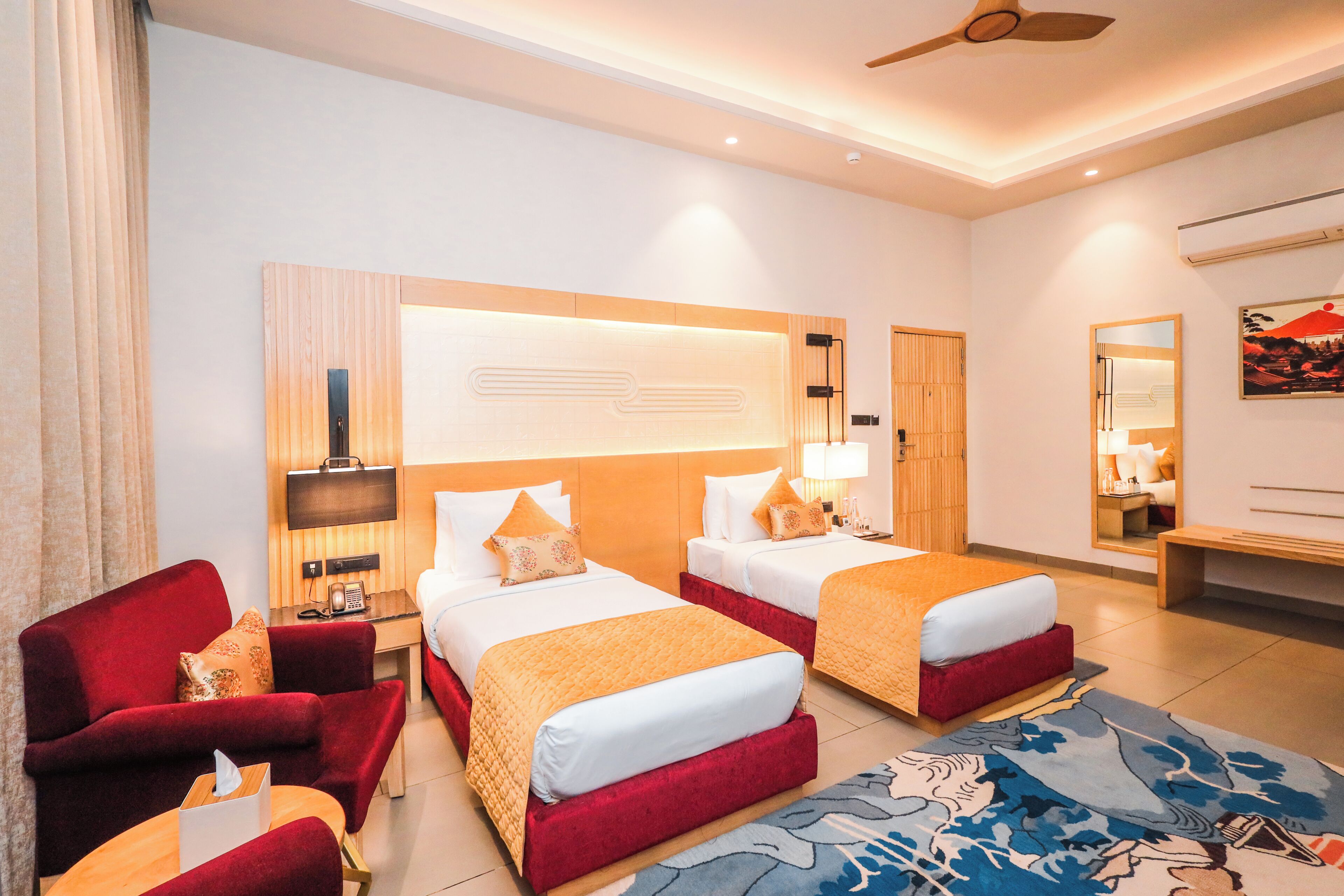 A hotel room with two beds, a red armchair, a small table, a ceiling fan, and a large mirror.