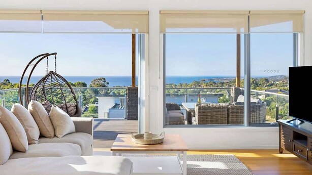Un salon moderne avec un canapé, un fauteuil suspendu, un téléviseur et de grandes fenêtres offrant une vue sur la mer et le paysage urbain.