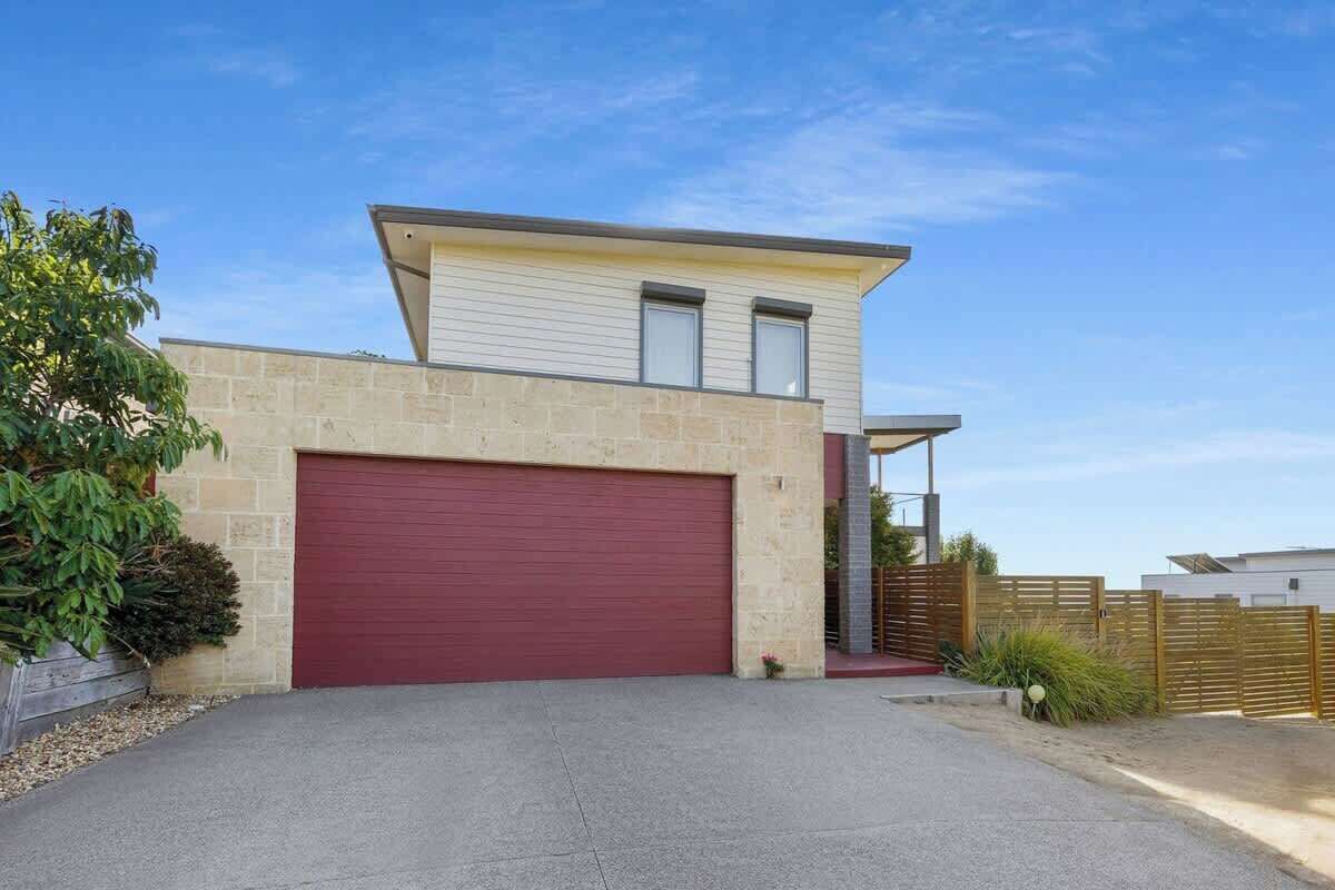 A modern two-story house with a red garage door, stone exterior, and a small front yard with a wooden fence and greenery.