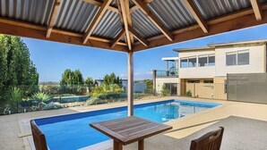 Un espace piscine extérieur abrité, avec une table et des chaises en bois, une maison à deux étages en arrière-plan et un ciel bleu dégagé.