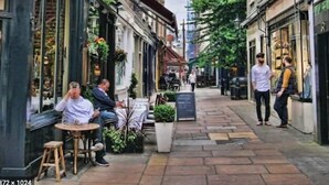 A narrow street with buildings on either side, outdoor seating for a cafe, and a few pedestrians.