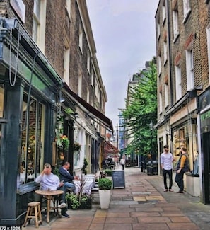Uma rua estreita com edifícios em ambos os lados, mesas ao ar livre para um café e alguns pedestres.