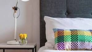 A neatly made bed with a grey upholstered headboard, a white pillow, and a colorful patterned pillow. A white bedside table with a lamp and a vase of yellow flowers is visible.