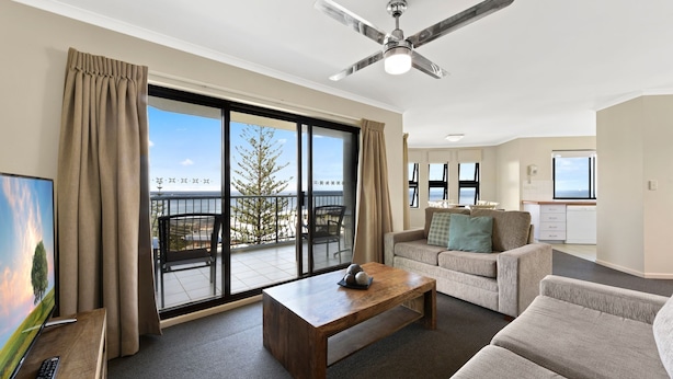A spacious living room with a large sliding glass door leading to a balcony, a ceiling fan, a wooden coffee table, and a sofa. - Gena Apartments Unit 13 Kings Beach QLD (Kings Beach)