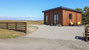 A wooden cabin with a gravel driveway, a wooden fence, and a view of the landscape. - The Wheelhouse (Isle of North Uist)