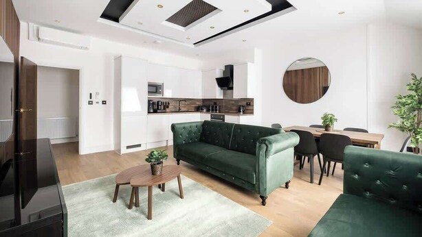 Ein modernes Wohnzimmer mit einem grünen Sofa, einem Holztisch und einem Fernseher. Im Hintergrund befindet sich eine Küche mit einer Mikrowelle sowie ein Essbereich mit Tisch und Stühlen.