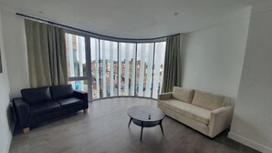 A modern living room with a black and beige sofa set, a round coffee table, and large floor-to-ceiling windows offering a city view.