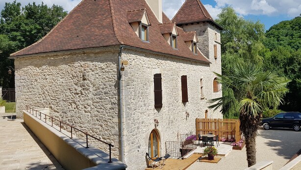 A stone building with a red-tiled roof, a wooden deck with outdoor seating, and a palm tree. - Moulin Proche Rivière Dans la Campagne Lotoise (Saint-Denis-Catus)