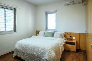 A bedroom with a bed, two windows, a nightstand, and a wooden wardrobe.