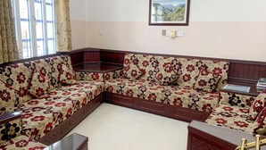 A corner sofa with floral upholstery, a small round table, and a framed picture on the wall. - Super OYO 152 Manam 1 Hotel Apartment (Muscat)