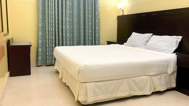 A hotel room with a large bed, a wooden headboard, a nightstand, a lamp, and a window with curtains. - Super OYO 152 Manam 1 Hotel Apartment (Muscat)