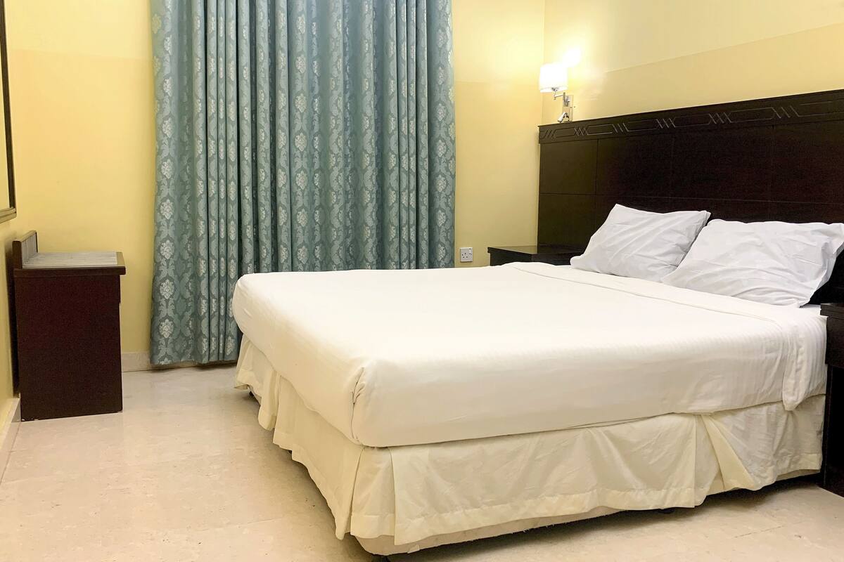 A hotel room with a large bed, a wooden headboard, a nightstand, a lamp, and a window with curtains.