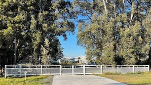 Une allée pavée mène à une zone délimitée par un portail et une clôture blanche, le tout étant entouré d’arbres et surplombé par un ciel dégagé.