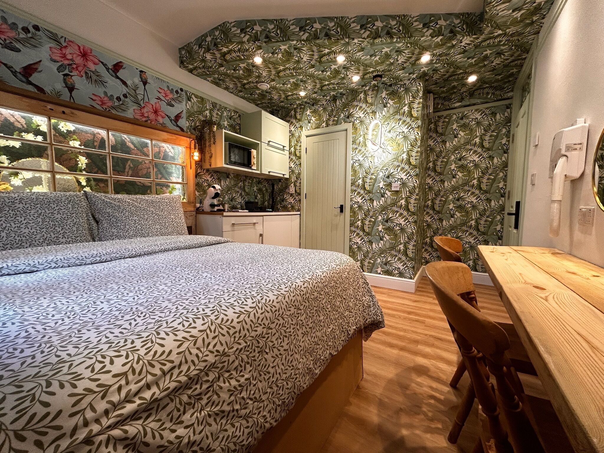 Un dormitorio con cama, un escritorio de madera y una silla. Se ve una ventana con vistas a árboles.