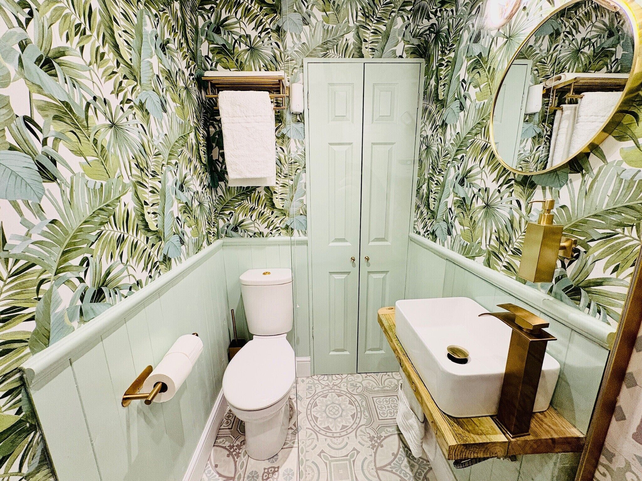 A bathroom with a toilet, sink, and mirror, surrounded by tropical wallpaper.