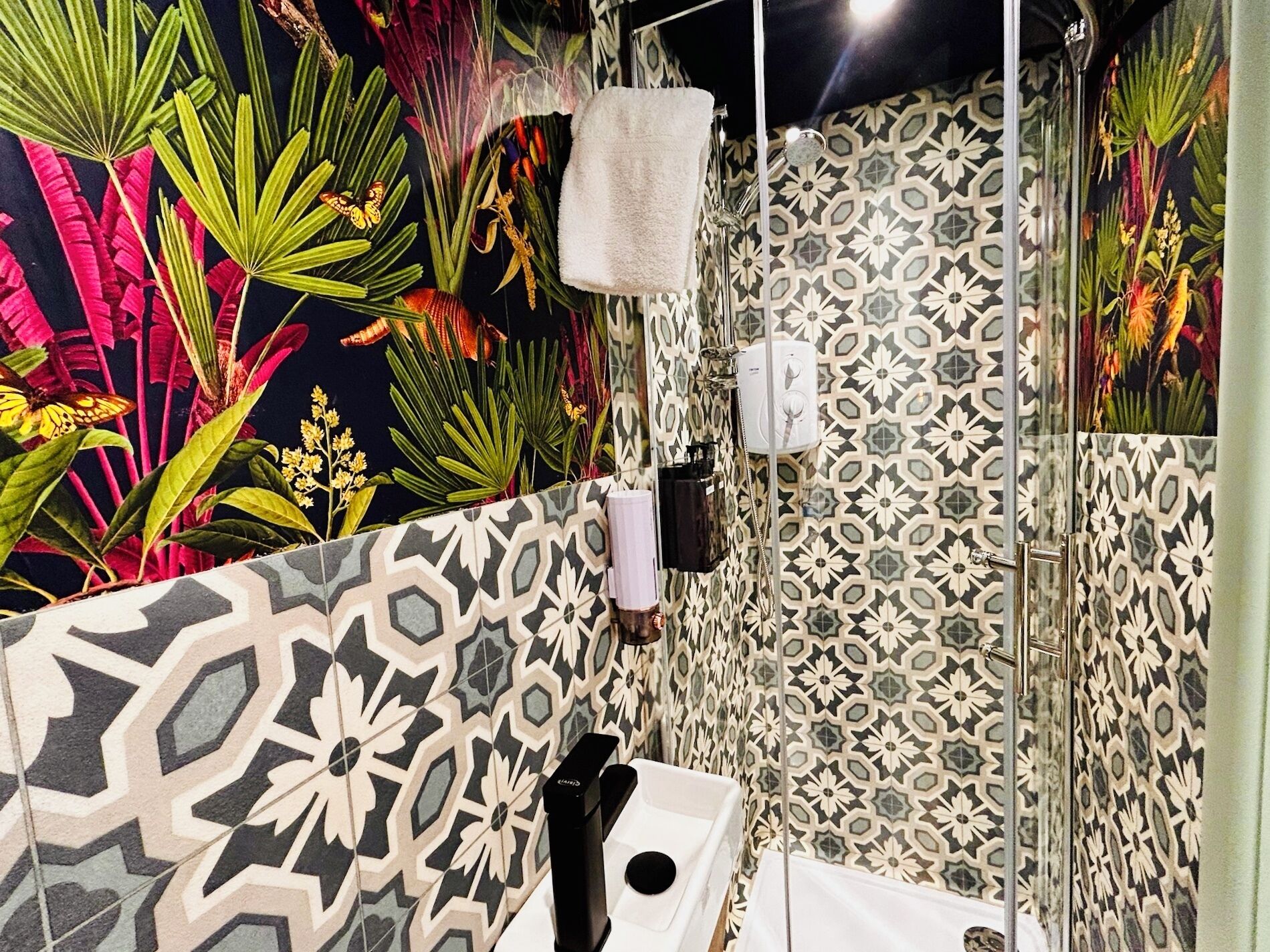 A bathroom with a shower, a sink, and a toilet. The walls are decorated with a tropical mural and patterned tiles.