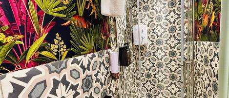 A bathroom with a shower, a sink, and a toilet. The walls are decorated with a tropical mural and patterned tiles.