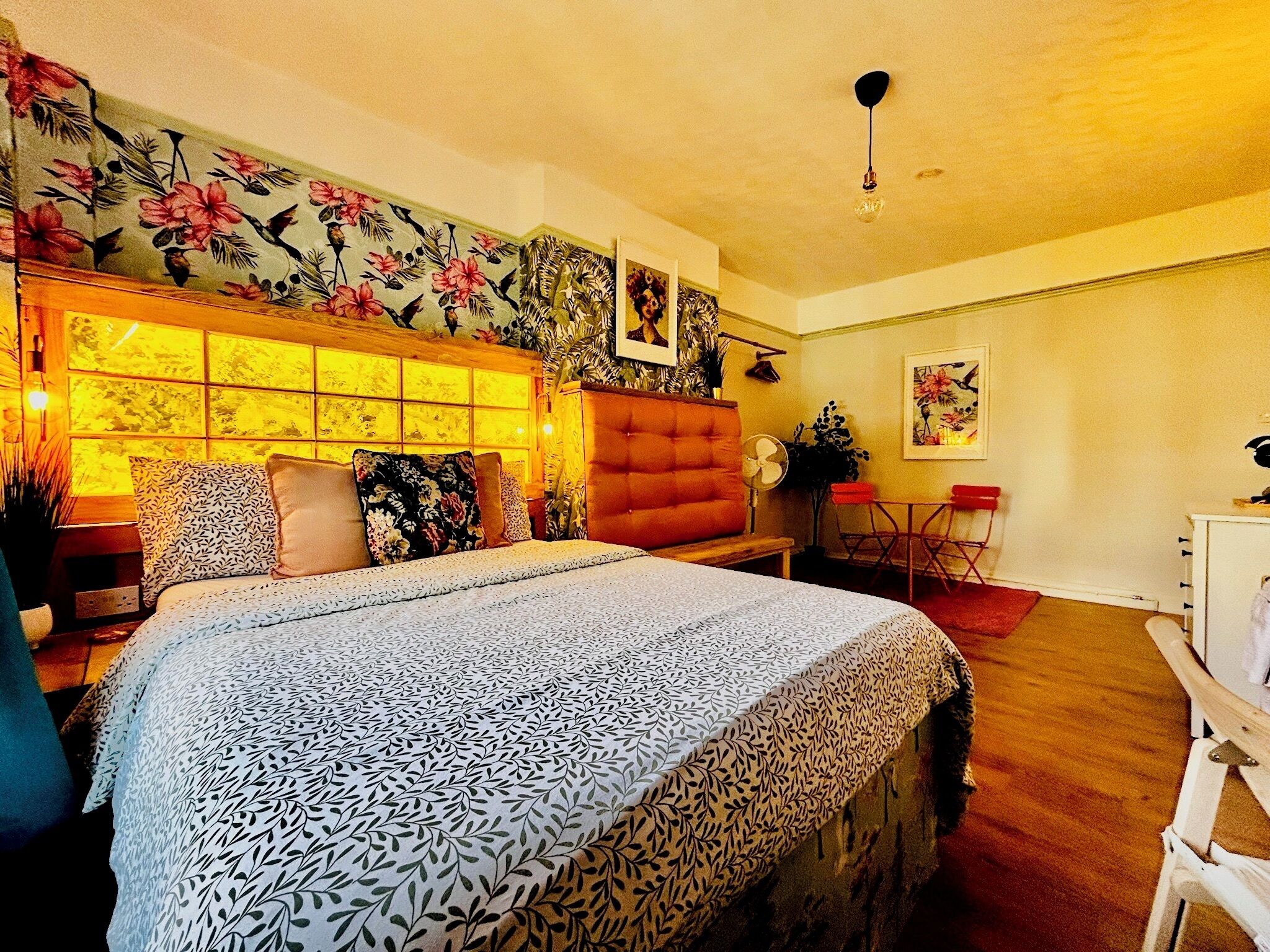 A bedroom with a large bed, a wooden headboard, a window with a floral view, a small table, and a chair.