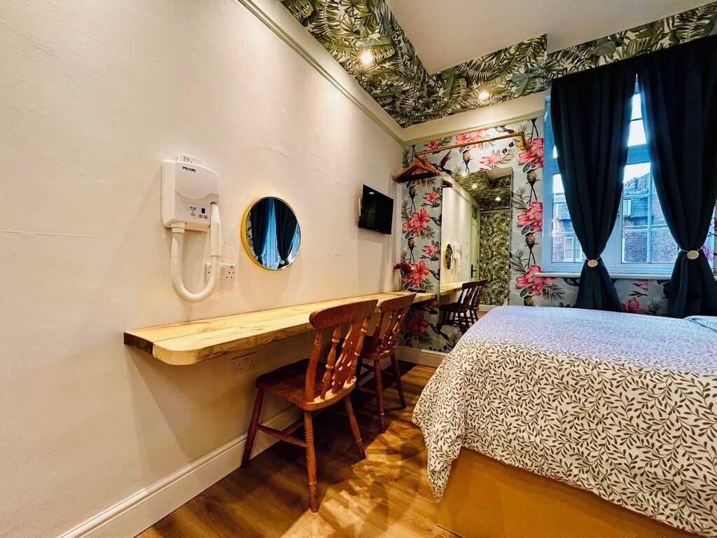 A room with a bed, a desk, and chairs. There is a mirror, a phone, and a window with curtains.