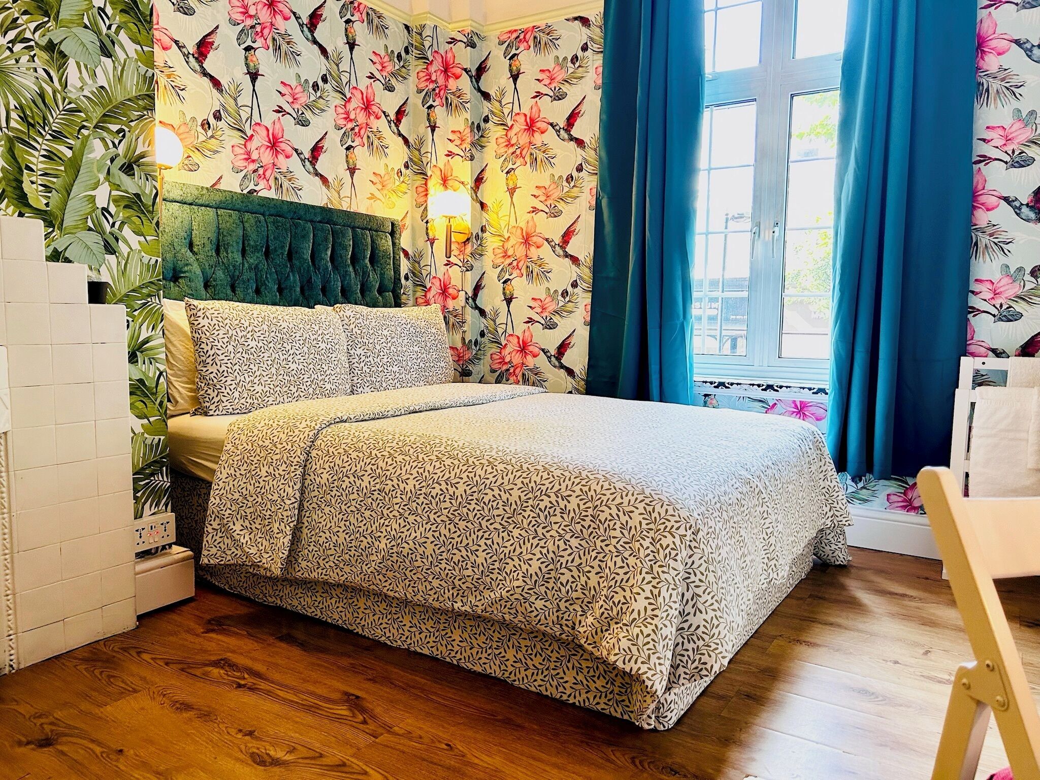 Een slaapkamer met bloemenbehang, een groen gestoffeerd hoofdeinde, een bed met beddengoed met een patroon, en een raam met blauwe gordijnen.